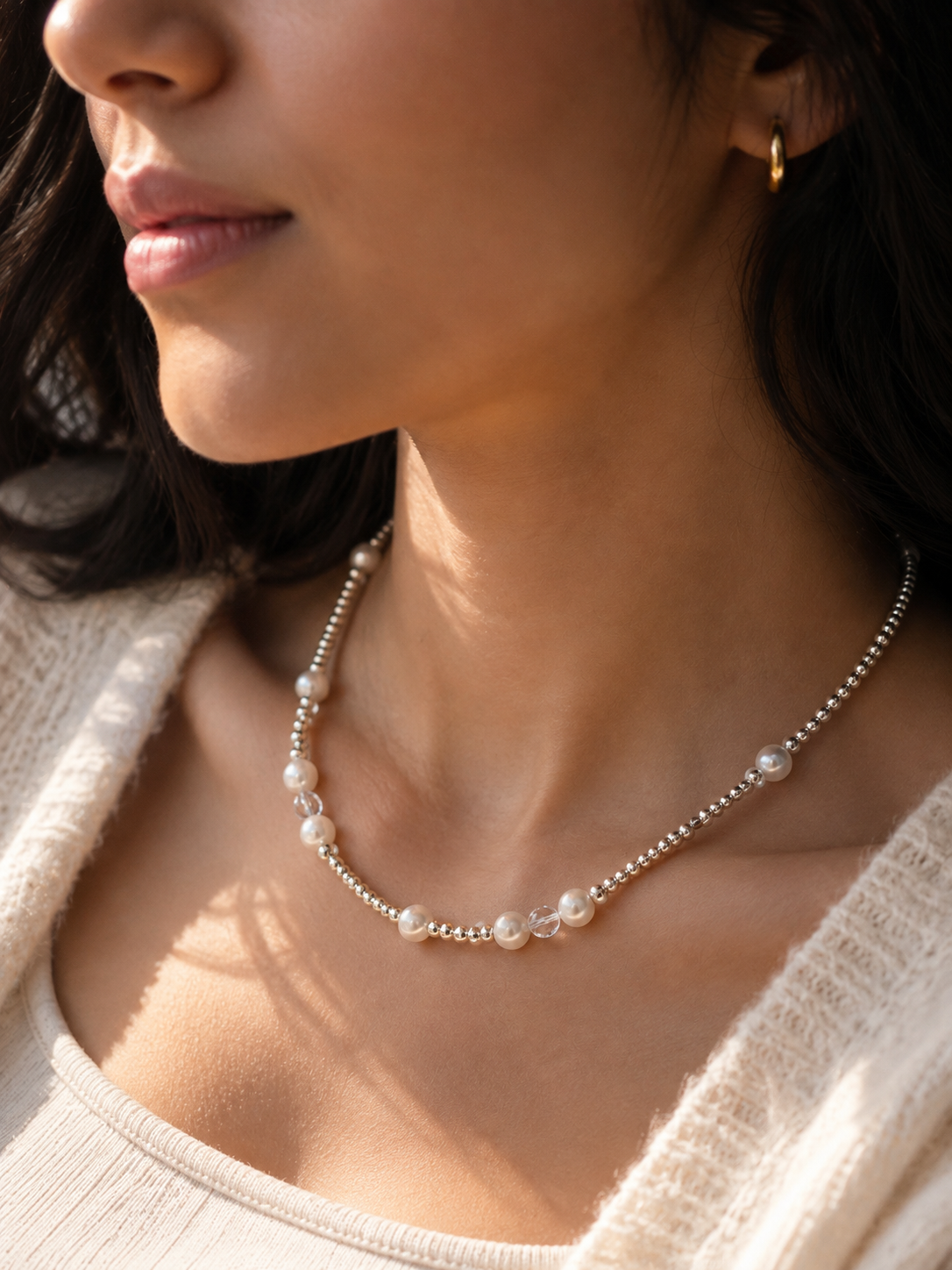 A close-up lifestyle shot of a woman wearing a delicate silver-toned beaded necklace with white pearl accents, styled with a small gold hoop earring.