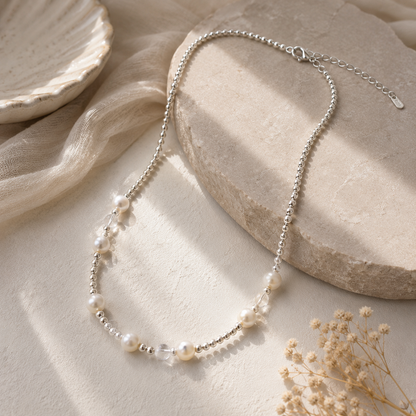 A delicate silver-toned beaded necklace with small white pearls and a clear faceted bead, styled on a textured beige surface with draped fabric and dried flowers.