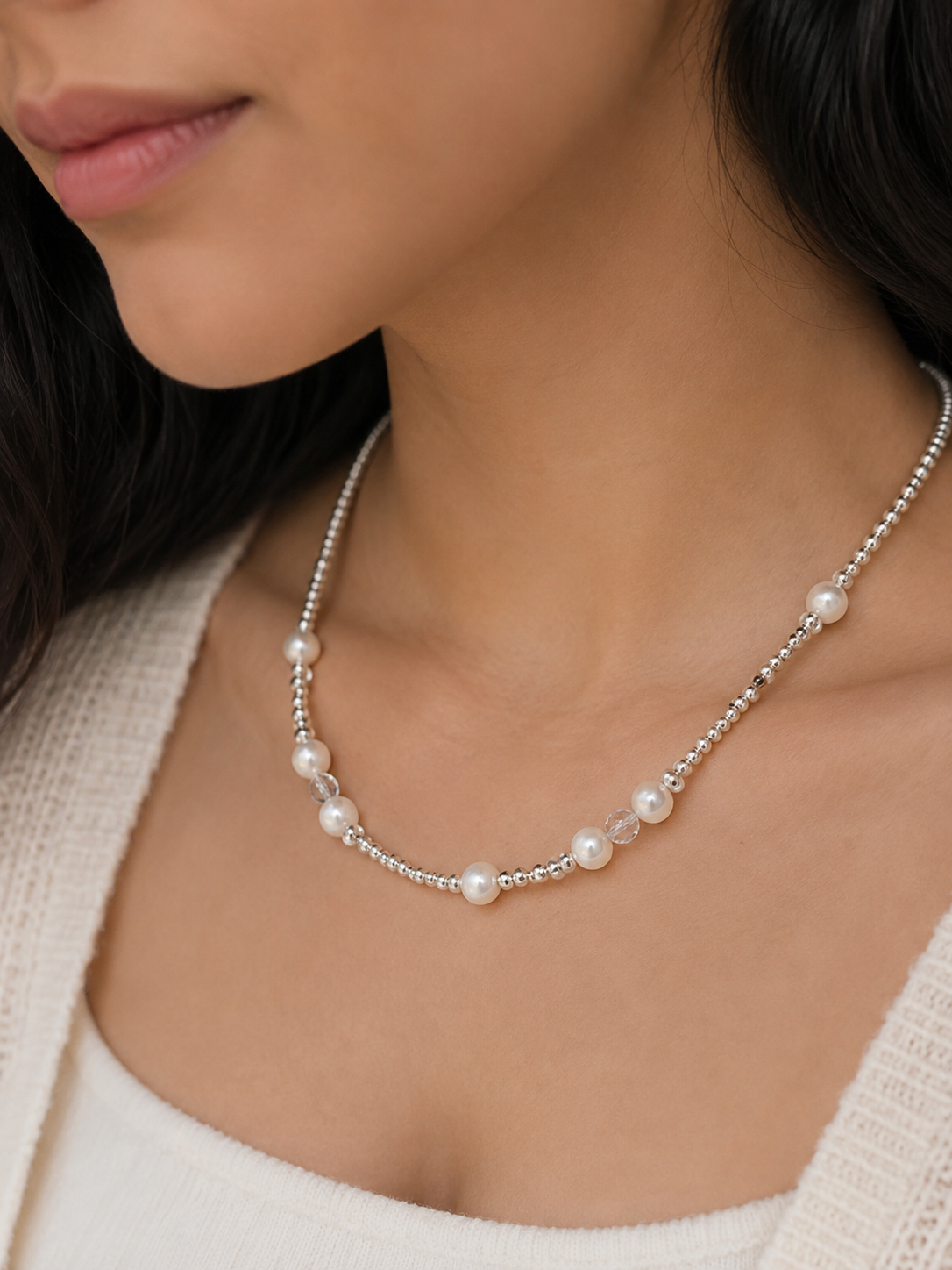 A close-up of a woman wearing a delicate silver beaded necklace with small white pearls spaced along the front.