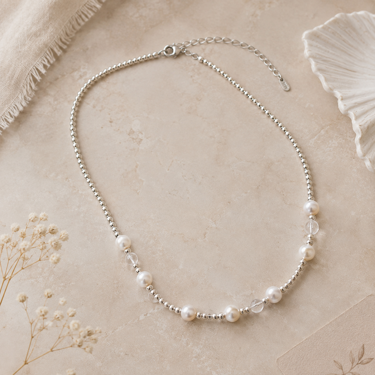 A delicate silver beaded necklace with scattered white freshwater pearls and clear faceted beads, arranged on a textured beige surface with dried floral accents.