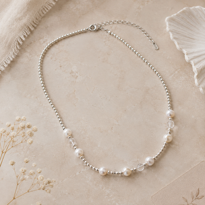 A delicate silver beaded necklace with scattered white freshwater pearls and clear faceted beads, arranged on a textured beige surface with dried floral accents.