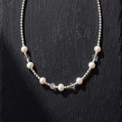 A silver beaded necklace with white pearl-like beads and clear faceted beads, displayed on a dark textured slate background.