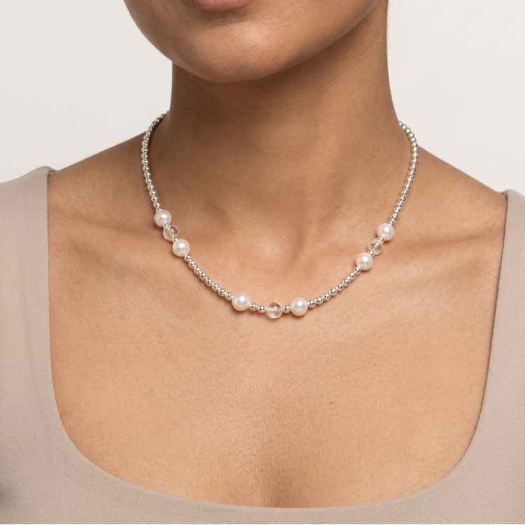 A woman with dark hair pulled back wears a delicate silver chain necklace with small white pearls and a central silver bead, paired with a beige scoop-neck top against a light gray background.