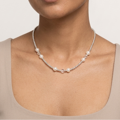 A woman with dark hair pulled back wears a delicate silver chain necklace with small white pearls and a central silver bead, paired with a beige scoop-neck top against a light gray background.