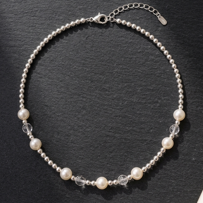 A delicate silver beaded necklace with white pearl accents and clear faceted crystal beads, displayed on a dark slate surface with a cream satin fabric corner.