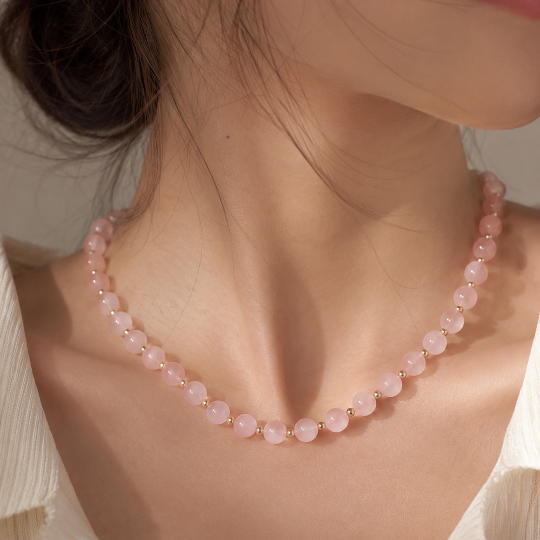 A close-up of a woman wearing a delicate pink beaded necklace with small gold spacer accents, styled with a cream pleated blouse.