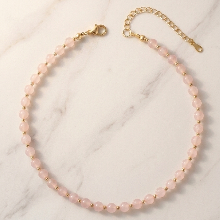 A delicate necklace made of small pale pink beads with gold-tone metal findings, arranged in a circle on a white marble surface.