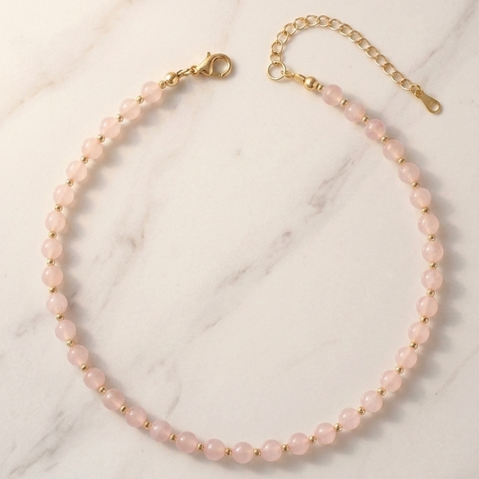 A delicate necklace made of small pale pink beads with gold-tone metal findings, arranged in a circle on a white marble surface.