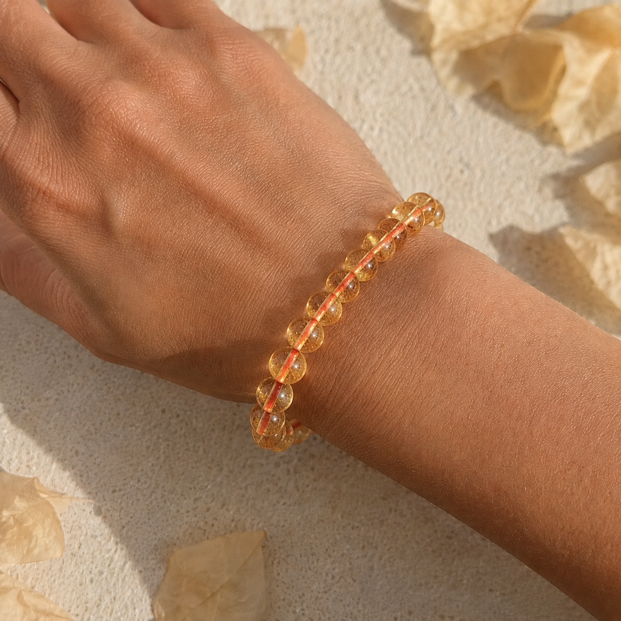 Hand wearing a citrine beaded bracelet on a textured beige surface with dried leaves.