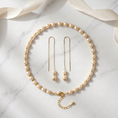 A pearl and gold jewelry set arranged on a white marble surface with a cream satin ribbon. The set includes a pearl necklace with gold spacer beads and a gold clasp, matching drop earrings with gold posts and pearl drops, and a small gold chain extender.