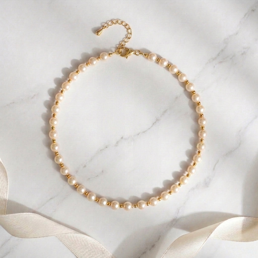 A delicate pearl necklace with gold spacer beads and a gold clasp, arranged in a circle on a white marble surface with a cream satin ribbon in the background.