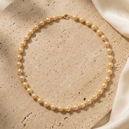A delicate double-strand pearl necklace with a gold clasp, arranged in a circle on a textured beige stone slab with draped white silk fabric.