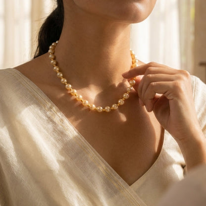 A woman wearing a delicate gold pearl necklace, adjusting it with her hand. She is wearing a cream-colored linen top in a softly lit room with warm sunlight and curtains.