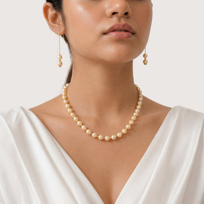 A close-up of a woman wearing a pearl necklace and matching drop earrings with gold-toned settings, against a clean light beige background.