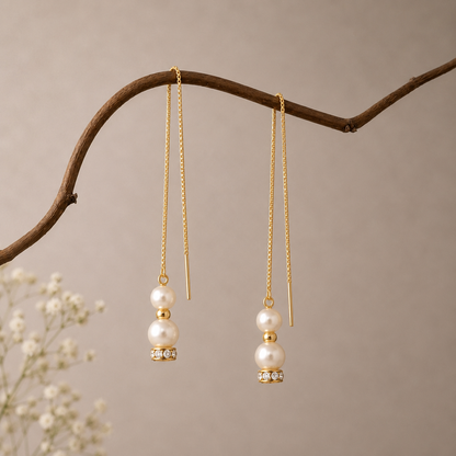 A pair of gold dangle earrings with white pearls and small clear rhinestones, hanging from a curved brown branch against a soft beige background.
