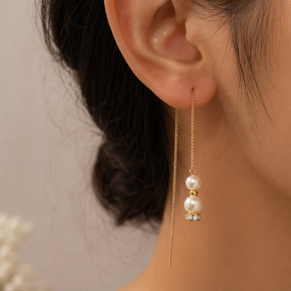 A close-up of a person's ear wearing a delicate gold dangle earring with two white pearl beads and a small clear crystal accent.