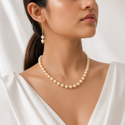 A close-up of a woman wearing a pearl necklace and matching drop earrings with gold-toned hooks, styled with a white satin top against a soft white background.