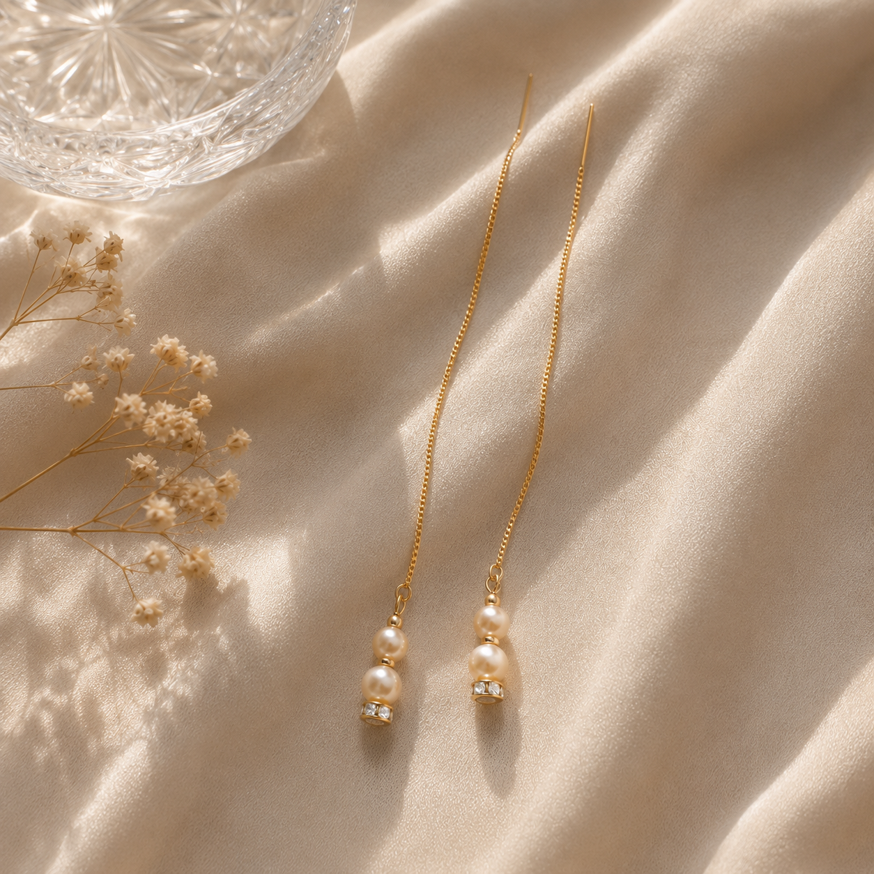 A pair of long gold chain earrings with double pearl drops and small clear crystal accents, arranged on draped cream fabric with dried flowers and a glass bowl nearby.