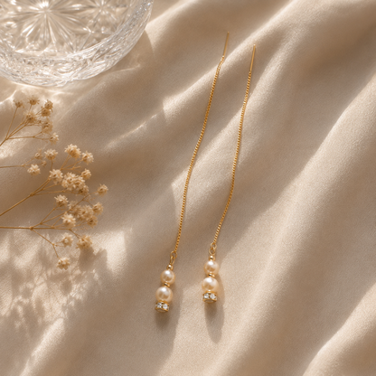 A pair of long gold chain earrings with double pearl drops and small clear crystal accents, arranged on draped cream fabric with dried flowers and a glass bowl nearby.