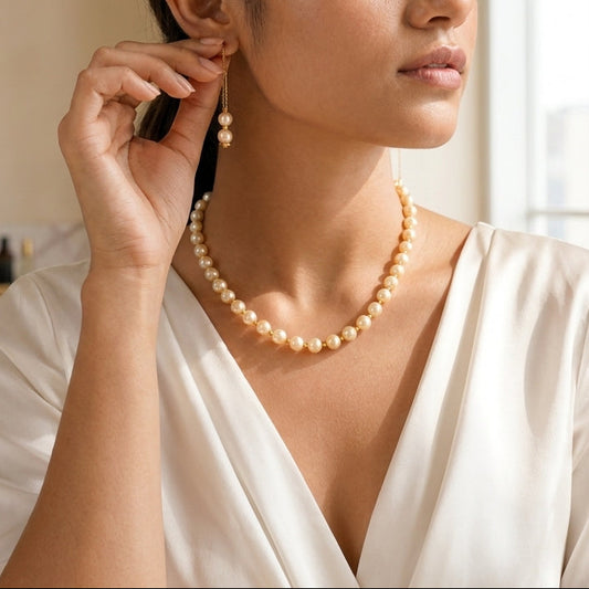 A woman wearing a pearl necklace and matching drop earrings, shown in a softly lit indoor setting. She is wearing a white satin V-neck top and looking to the side while adjusting or wearing the earring with her hand near her ear.