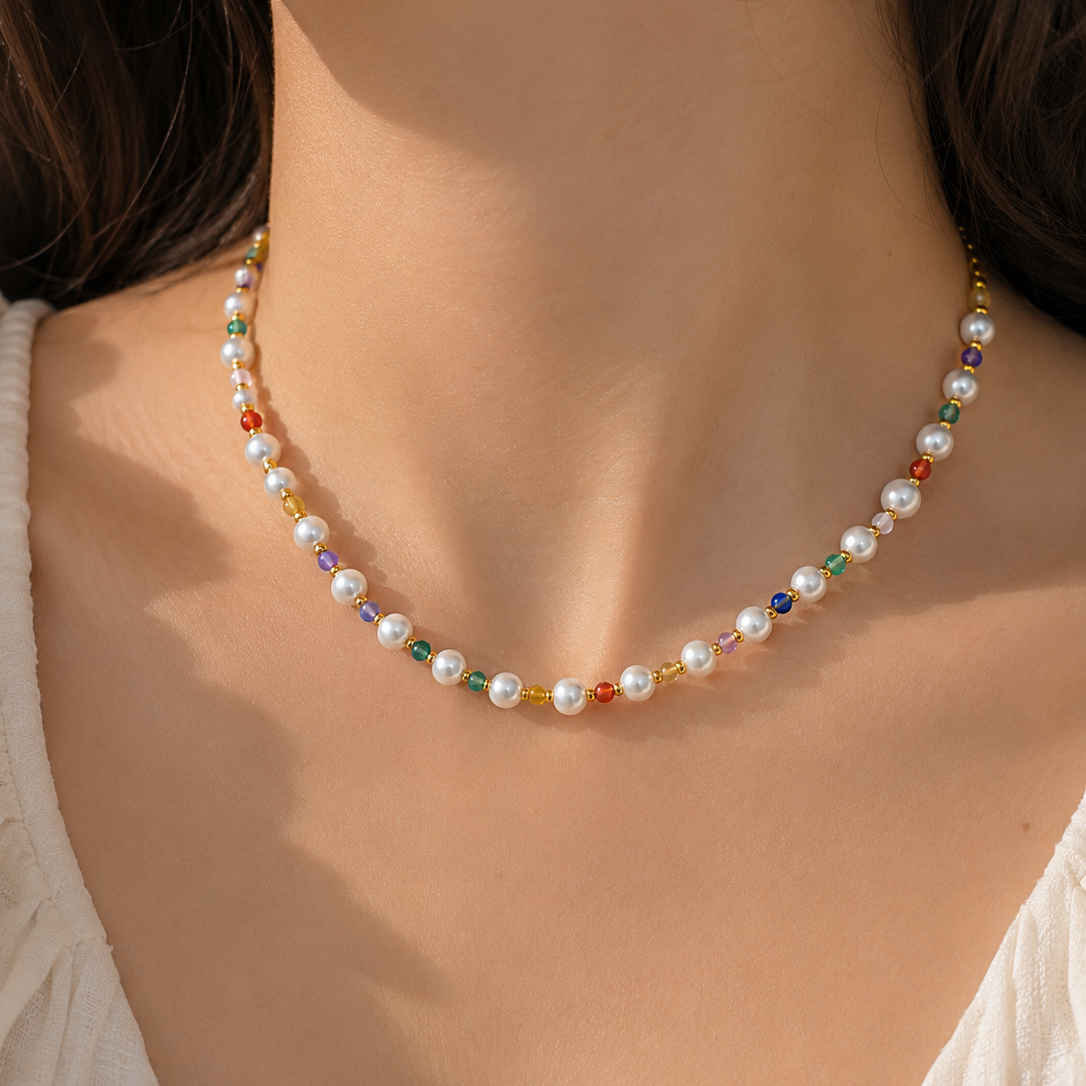A close-up of a model's neck and chest wearing a delicate necklace made of white pearls and small multicolored beads separated by gold spacer beads, paired with a white top.