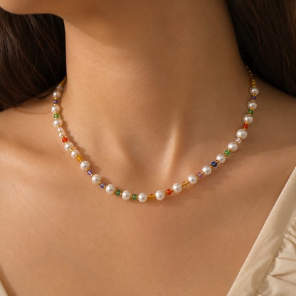 A close-up of a model's neck and upper chest wearing a delicate necklace made of white freshwater pearls separated by small multicolored faceted beads.