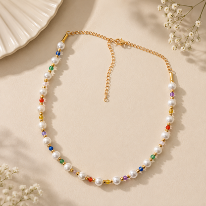 A gold chain necklace with white faux pearls and small colorful gemstone beads, arranged in a loop on a warm beige surface with decorative flowers and a ceramic dish in the corners.