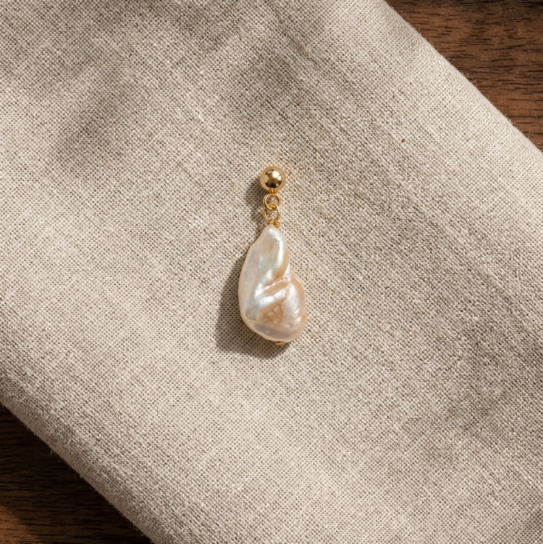 A single teardrop-shaped freshwater pearl earring with a gold post and bead, resting on a textured beige linen cloth over a dark wood surface.