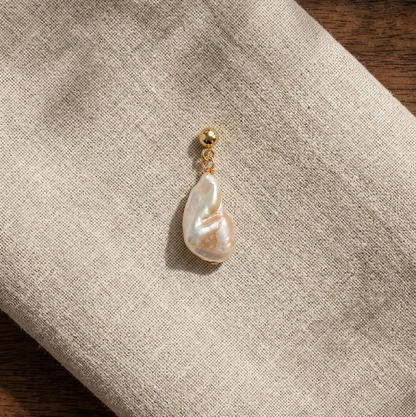 A single teardrop-shaped freshwater pearl earring with a gold post and bead, resting on a textured beige linen cloth over a dark wood surface.