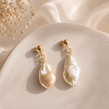 A pair of gold drop earrings with irregular baroque pearl-like pendants, displayed on a beige fabric surface with dried flowers in the corner.